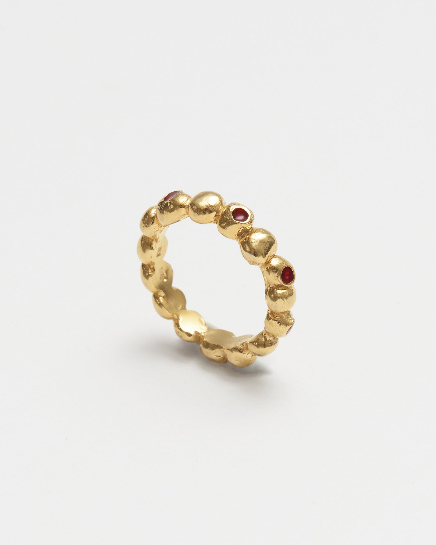 A textured gold ring with small red gemstone accents, photographed on a clean light gray background.