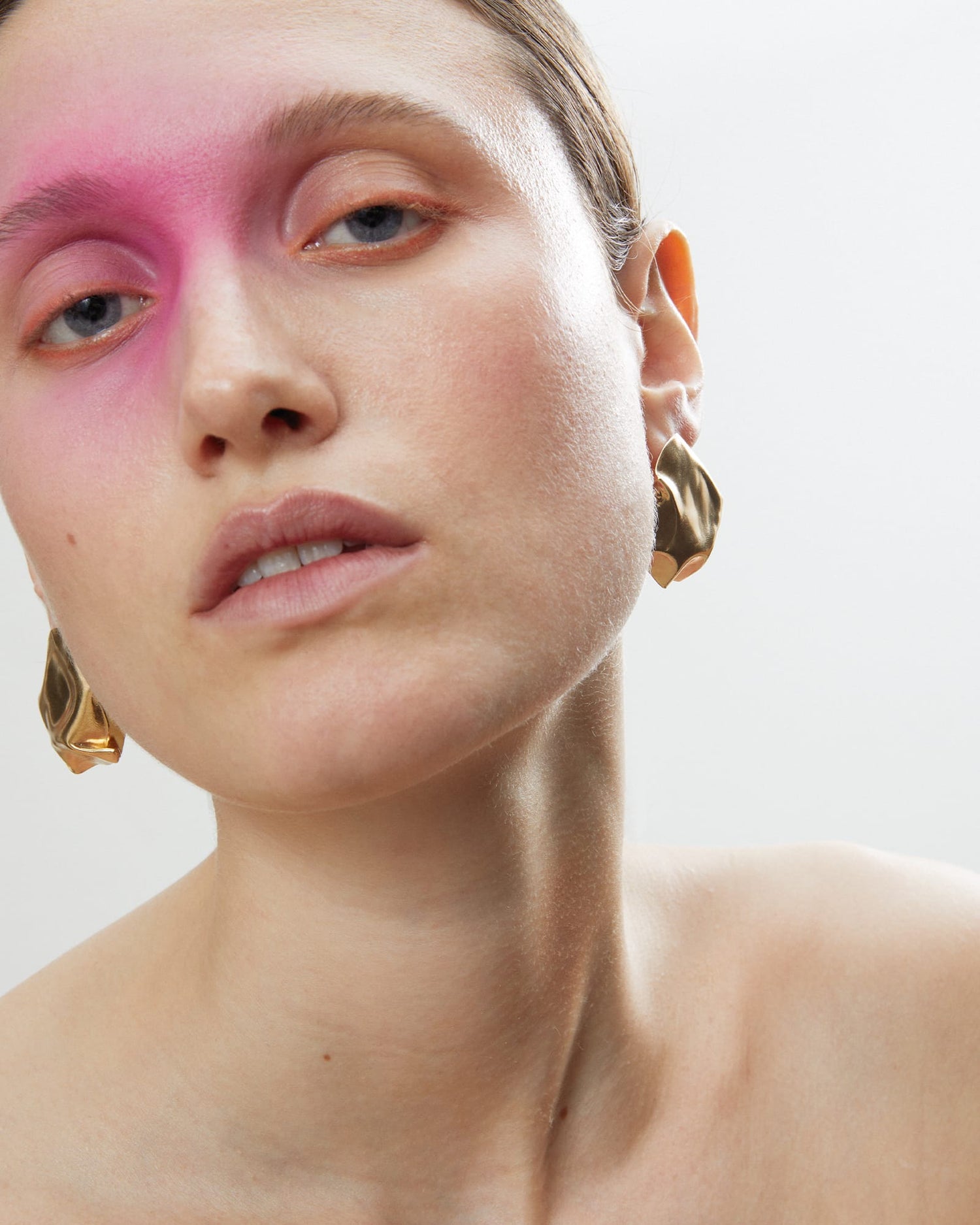 Close-up of a model wearing LMJ Blow Earrings in gold, styled with pink makeup and slicked-back hair.