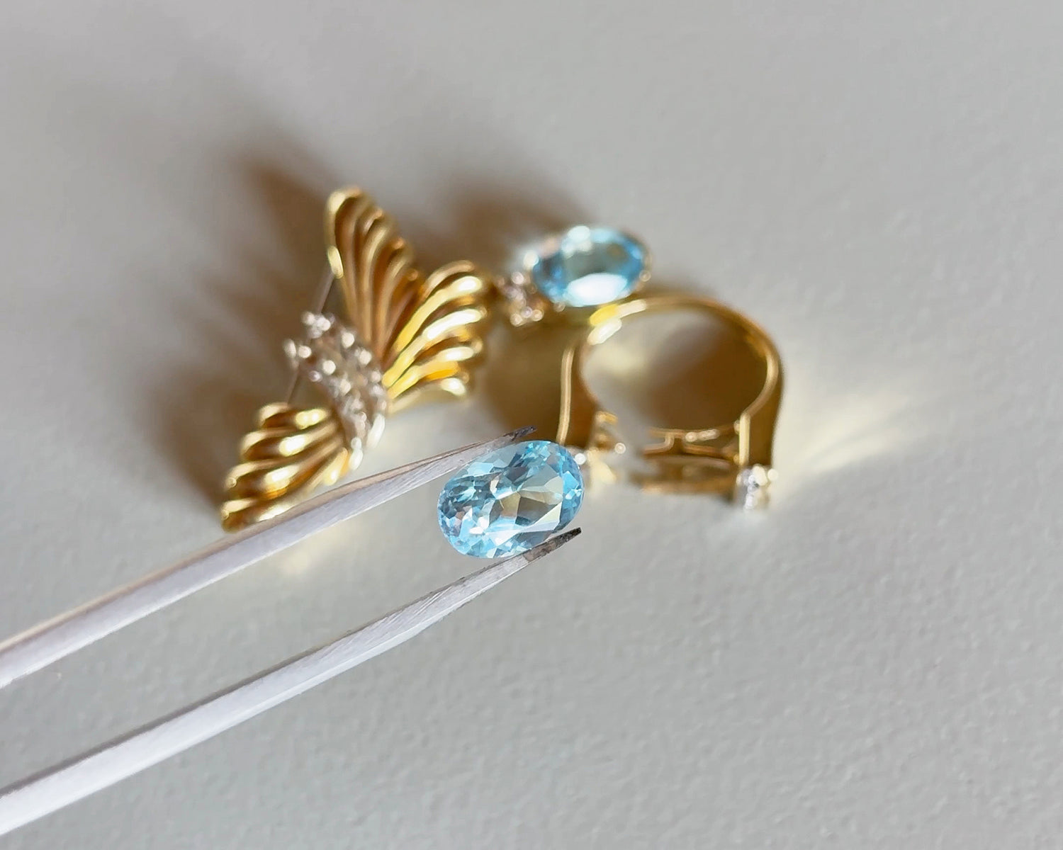 Close-up of an aquamarine being held by tweezers and other rings and brooch on the background.