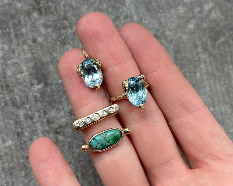 Three gold rings with gemstones and a pendant with an aquamarine on a hand against a grey background