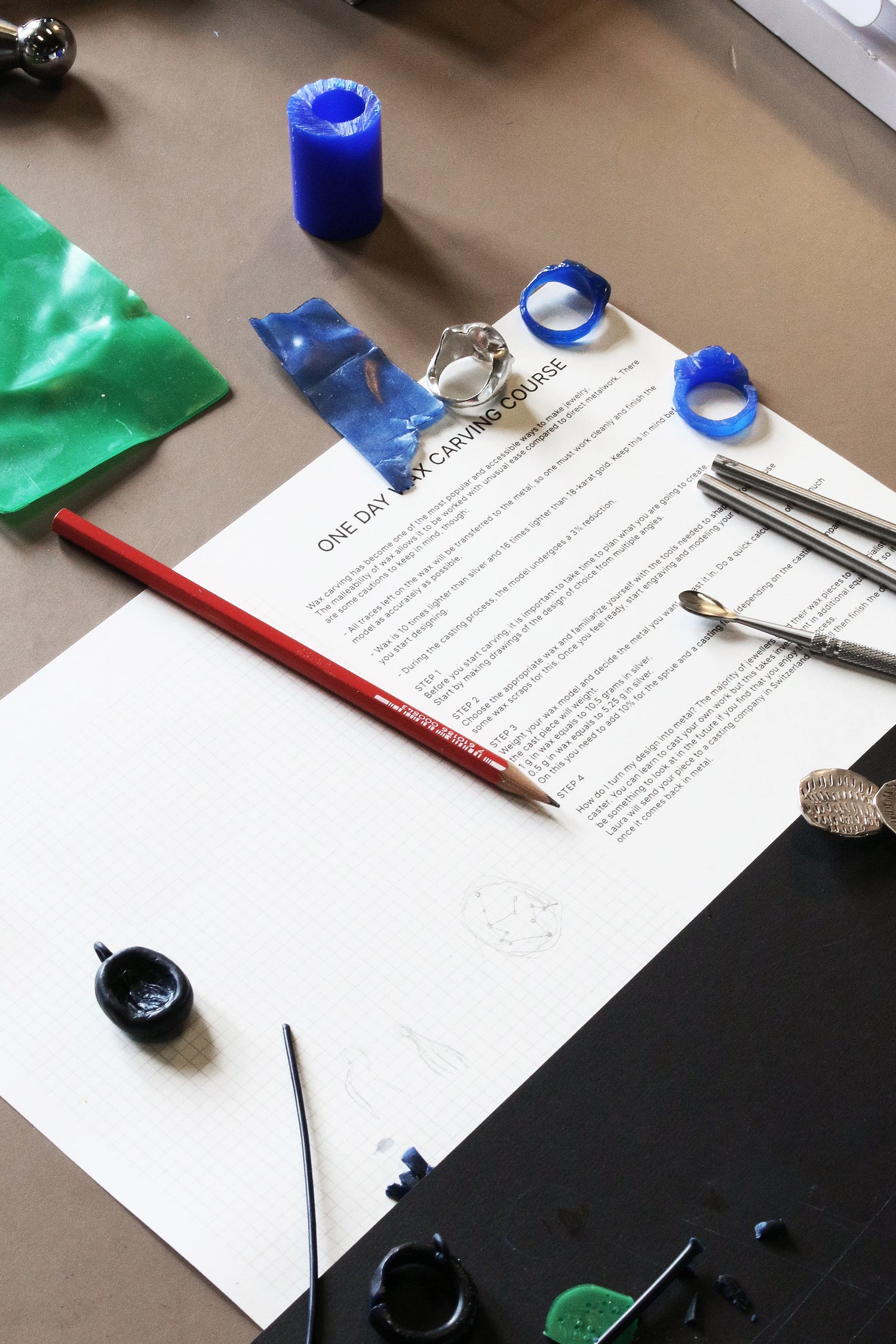 Document on a desk with art supplies including paint, brushes, during the during the LMJ wax carving jewelry workshop.